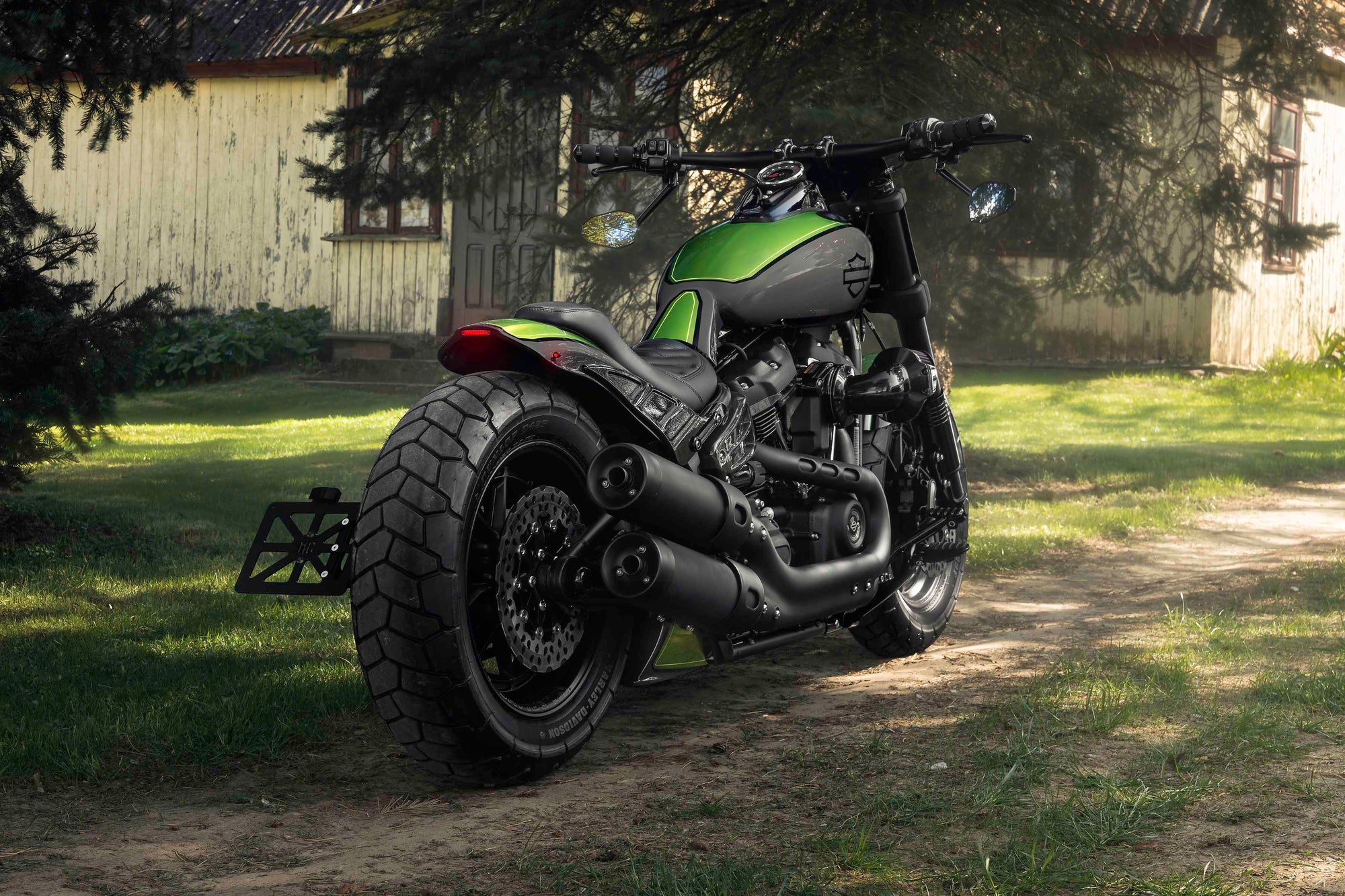 Modified Harley Davidson Fat Bob motorcycle with Killer Custom parts from the rear with a country house and some trees in the background